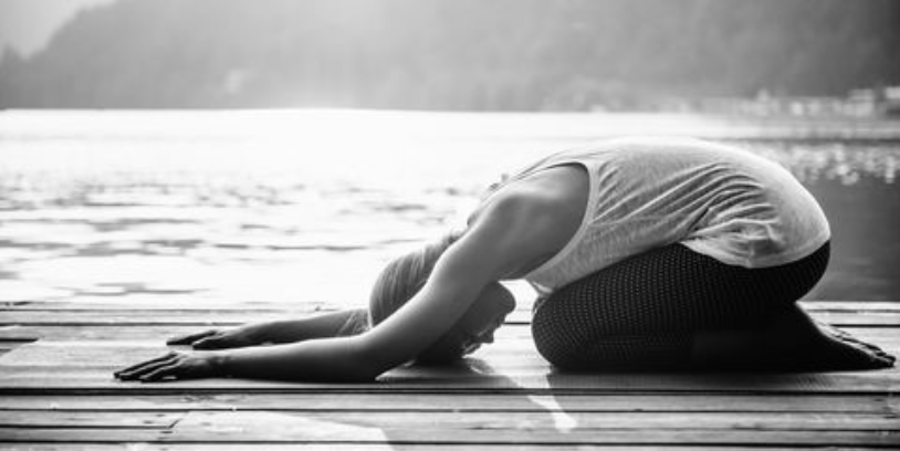 Vrouw die yoga doet op een houten ondergrond naast een meer met bergen op de achtergrond.